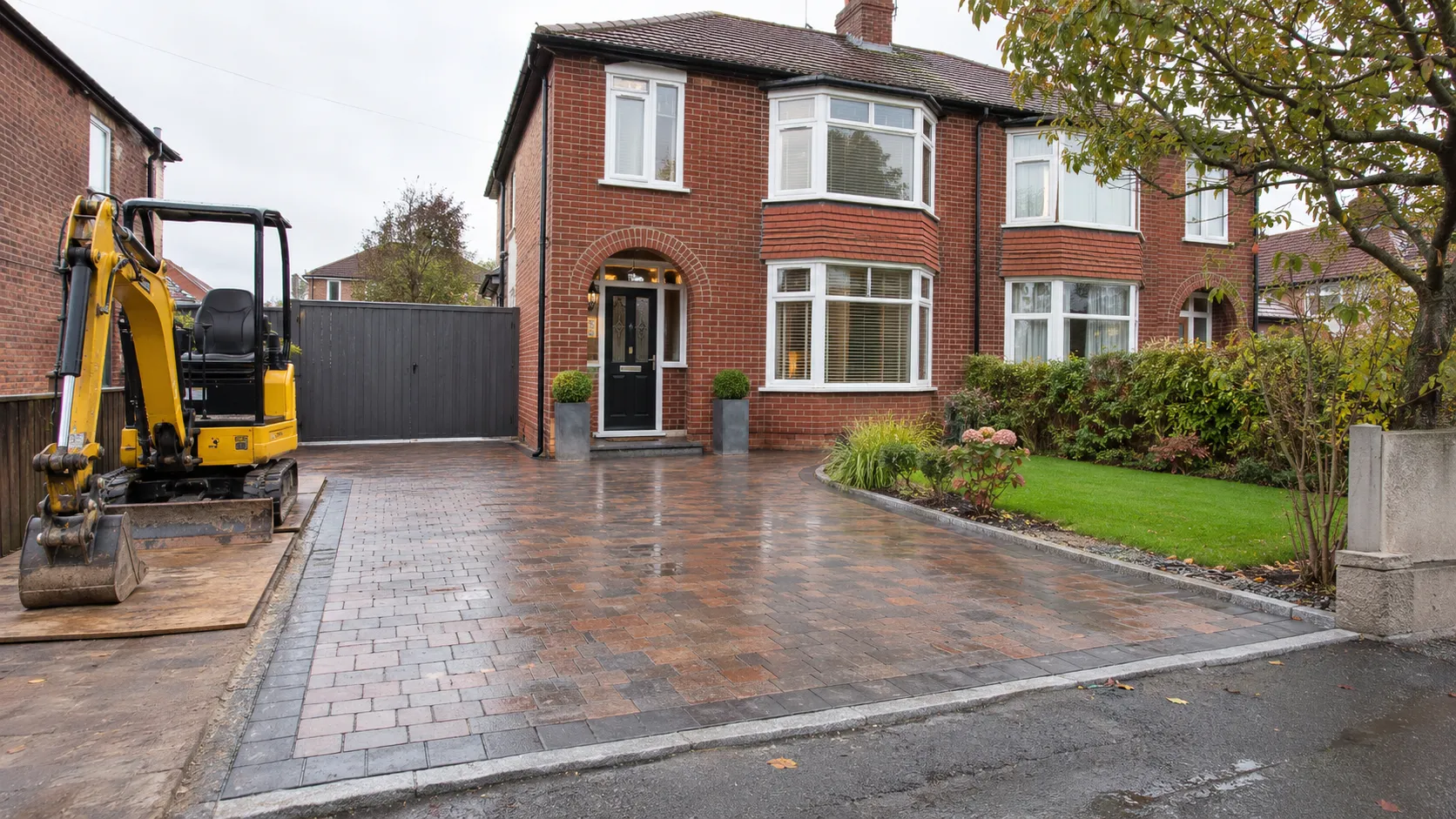 A new block paving driveway with granite edging in front of a red brick semi, with a mini digger on boards