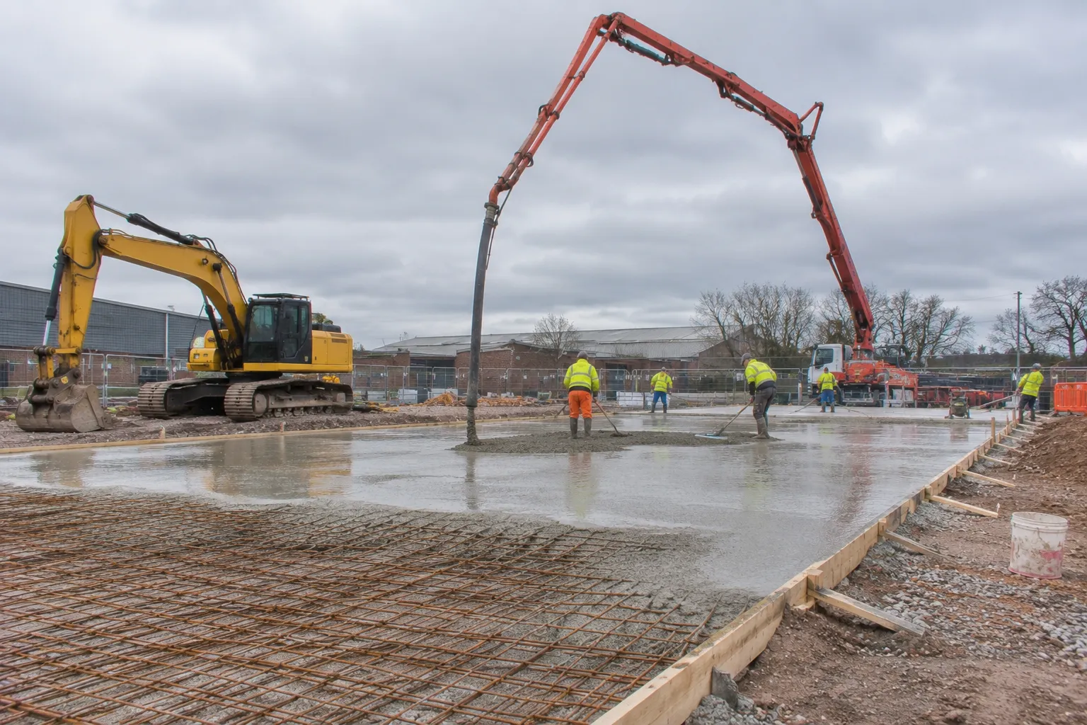 A reinforced concrete slab being poured from a concrete pump on a commercial site, with an excavator to one side
